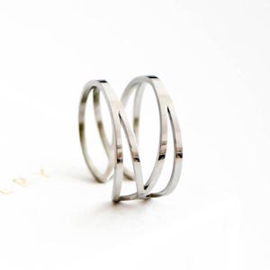 Peut inclure: Une bague argentée au design minimaliste. La bague comporte trois anneaux, dont deux arrondis et un plat. La bague est posée sur un fond blanc, avec le nom de la marque "LVN JEWELRY" visible.