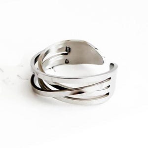 Puede incluir: Un anillo plateado con un diseño único e entrelazado. El anillo presenta múltiples bandas que se cruzan, creando un aspecto intrincado y moderno. El anillo está hecho de un metal brillante y reflectante, y está sobre un fondo blanco.