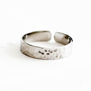 Puede incluir: Un anillo ajustable plateado con textura martillada. El anillo está abierto en la parte superior y descansa sobre una superficie blanca. El anillo es una joya.