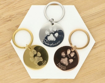 May include: Three silver, gold, and rose gold keychains with paw print engravings. Each keychain features a round tag with a paw print and a name engraved on it. The names are Sundance, Frankie, and Indie.