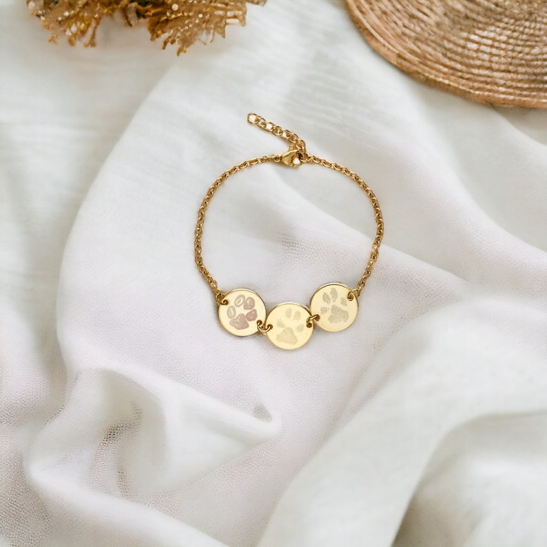 Puede incluir: Un brazalete dorado con tres colgantes circulares, cada uno con una huella de pata grabada. El brazalete est&aacute; sobre una tela blanca texturizada. La cadena y los colgantes tienen un acabado pulido. Las huellas son de color rosa p&aacute;lido.