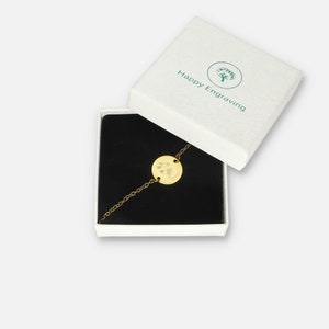 Puede incluir: Una pulsera dorada con un colgante circular, presentada en una caja de regalo blanca abierta. El colgante est&aacute; grabado con un peque&ntilde;o dise&ntilde;o y el nombre "Chloe". La caja lleva la marca "Happy Engraving" y tiene un interior negro.