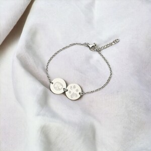 Puede incluir: Una pulsera plateada con dos colgantes circulares. Uno muestra una huella de pata, el otro una huella de nariz. La pulsera tiene una cadena fina y un cierre ajustable. El fondo es una tela blanca texturizada.