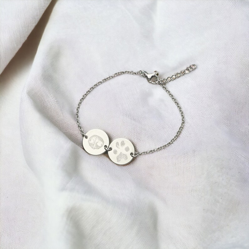 Puede incluir: Una pulsera plateada con dos colgantes circulares. Uno muestra una huella de pata, el otro una huella de nariz. La pulsera tiene una cadena fina y un cierre ajustable. El fondo es una tela blanca texturizada.