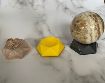Puede incluir: Tres soportes de exhibición geométricos hechos de resina. Los soportes tienen forma de diamante. Un soporte es amarillo, otro es marrón claro y el último es negro. El soporte marrón claro tiene un pequeño cuenco en el centro. El soporte negro tiene una esfera redonda marrón claro y blanca en la parte superior.
