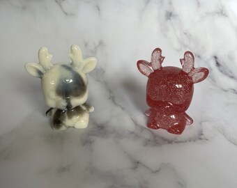 May include: Two small reindeer figurines, one white with black spots and the other pink with glitter.