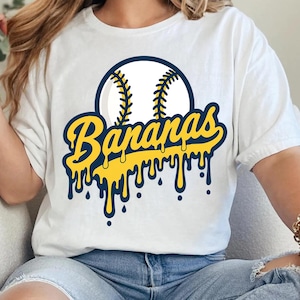 以下が含まれることがあります： 白い半袖Tシャツで、野球ボールのグラフィックの下に、濃い青色の縁取りが施された黄色い滴るフォントで「Bananas」という文字がデザインされています。このデザインはTシャツの胸の中央に配置されており、青いジーンズと合わせて着用されています。