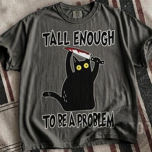 May include: A charcoal gray short-sleeve t-shirt featuring a graphic of a black cat with yellow eyes holding a knife above its head. The text "TALL ENOUGH TO BE A PROBLEM" is printed in white capital letters above and below the cat illustration.