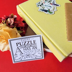 May include: A yellow hardcover journal with a floral and hummingbird sticker, a small brown notebook, and a sticker that reads "Every Puzzle Has An Answer" on a red surface. Decorative flowers are in the background.