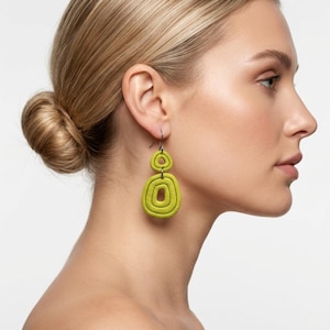 May include: Lime green, geometric drop earrings. The earrings feature a layered, circular design with a textured appearance. The earrings are suspended from a dark metal hook. The overall style is modern and bold.