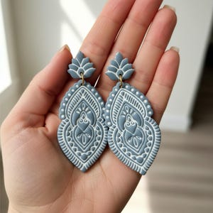 May include: A pair of gray and white patterned earrings. The earrings have a teardrop shape with intricate floral and dot designs. The earrings are held up by a gold-colored hook.