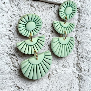 May include: Mint green statement earrings with a layered design. Each earring features a circular top piece and two half-circle pendants, all with a textured, fan-like pattern. Gold-colored hoops connect the pieces. The earrings are displayed against a textured, light gray background.