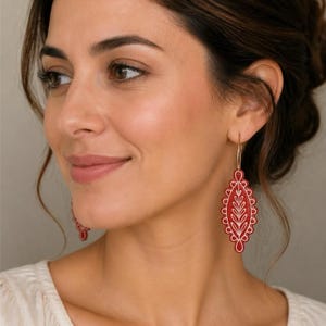 May include: A pair of dangling earrings featuring a red, elongated, scalloped-edge pendant with a white embroidered leaf pattern. The pendant is suspended from a thin gold-toned metal hoop wire that passes through the earlobe.