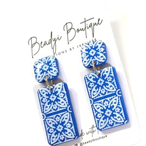 May include: Blue and white patterned earrings. The earrings have a square top and a rectangular bottom, connected by a gold-colored ring. The earrings feature a repeating white floral design on a blue background. The card behind the earrings reads "Beadyi Boutique".