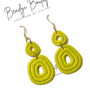 May include: A pair of lime green dangle earrings featuring two stacked, textured, organic-shaped loops suspended from gold-toned metal hooks. The earrings are displayed against a white card with the text Beadyi Boutique Creations by Ivelisse.