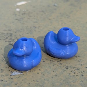 Puede incluir: Dos patos de goma azules. Los patos están sentados sobre una superficie marrón claro.