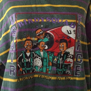 Puede incluir: Una camiseta de manga larga a rayas verdes y moradas con un estampado gráfico de un avión de dibujos animados y cuatro personajes de dibujos animados. El gráfico dice "US AIR FORCE" y "BIRDY SQUAD" con el texto "Always ready to fly" debajo del gráfico.