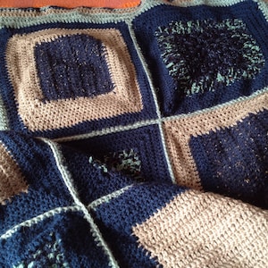 Può includere: Una coperta all'uncinetto fatta a mano nei toni blu navy, azzurro e beige. La coperta presenta un design patchwork con motivi quadrati e rettangolari. I bordi sono smerlati, aggiungendo un tocco decorativo. La coperta è piegata.