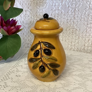 Peut inclure: Un pot en céramique jaune moutarde avec un couvercle assorti doté d'un petit bouton noir. L'avant du pot est orné d'un relief tridimensionnel représentant une branche d'olivier vert foncé avec des olives noires et des feuilles vertes.