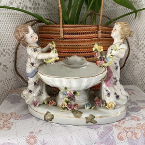 Puede incluir: Centro de mesa de porcelana vintage con dos figuras con vestidos florales que flanquean una pila. La base está adornada con flores y ribetes dorados. Las figuras sostienen cestas y pequeños pájaros amarillos. Sobre un mantel floral.
