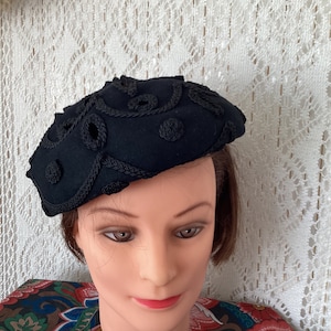 May include: A black felt beret with a decorative braided design. The beret has a rounded shape and is adorned with small black knots.