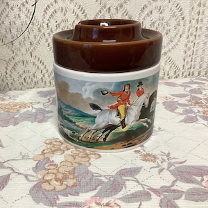 May include: A brown and white ceramic jar with a lid. The jar features a scene of two people riding horses in a field. The jar is approximately 15 cm tall.