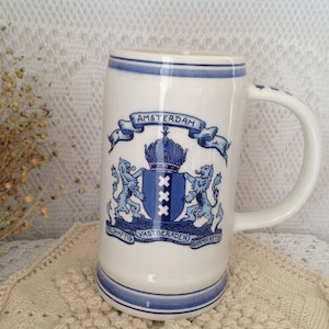 Peut inclure: Chope à bière en céramique blanche avec des détails bleus et les armoiries d'Amsterdam. La chope a une anse et des bandes bleues en haut et en bas. Les armoiries d'Amsterdam comprennent une couronne, des lions et le mot "Amsterdam".