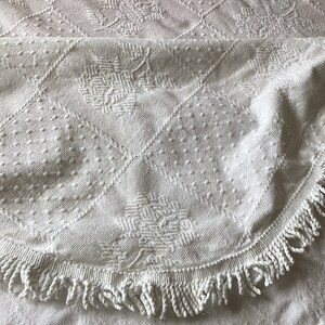 May include: White, textured textile with a floral and geometric pattern. The fabric features a diamond grid design with dotted and woven details. A fringed edge curves along the bottom, adding a decorative element.