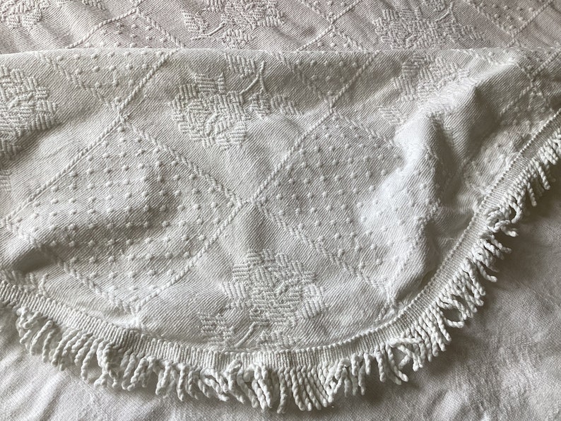 May include: White, textured textile with a floral and geometric pattern. The fabric features a diamond grid design with dotted and woven details. A fringed edge curves along the bottom, adding a decorative element.
