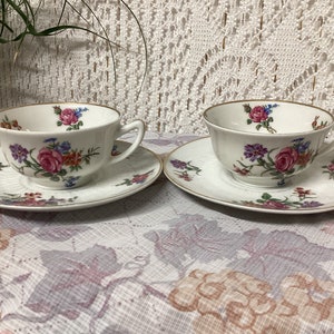 Peut inclure: Deux tasses à thé et soucoupes blanches avec un motif floral dans des tons de rose, rouge et bleu. Les tasses ont un bord doré et les soucoupes ont un design gaufré délicat.