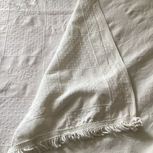 May include: White, textured tablecloth with a square pattern and floral designs. The tablecloth is folded over, revealing a fringe trim along the edge. The fabric appears to be cotton or linen.