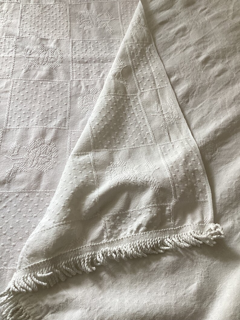 May include: White, textured tablecloth with a square pattern and floral designs. The tablecloth is folded over, revealing a fringe trim along the edge. The fabric appears to be cotton or linen.