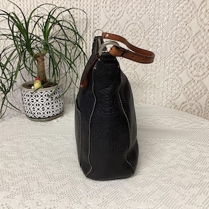 Pu&ograve; includere: Borsa a mano in pelle nera con texture martellata, cuciture bianche e manico in pelle marrone. La borsa ha una forma arrotondata e una tasca laterale. Il manico &egrave; fissato con anelli in metallo argentato.