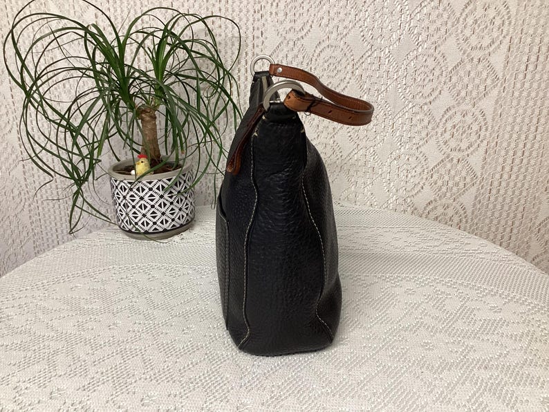 Pu&ograve; includere: Borsa a mano in pelle nera con texture martellata, cuciture bianche e manico in pelle marrone. La borsa ha una forma arrotondata e una tasca laterale. Il manico &egrave; fissato con anelli in metallo argentato.
