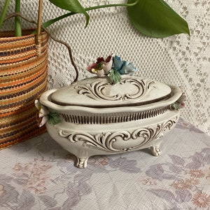 May include: A white ceramic trinket box with a floral design and a lid. The box has a decorative floral design on the lid and is decorated with three-dimensional flowers.