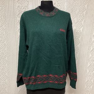 Op de afbeelding: Een groene trui met een rode en zwarte gepatroneerde rand rond de hals en onderkant. De trui heeft een klein rood logo met de tekst "Corduroy" op de voorkant.