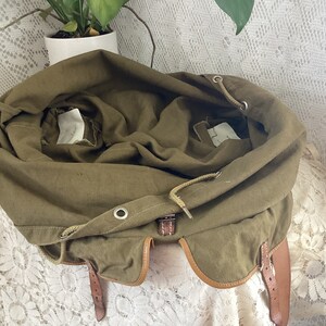 May include: A green canvas backpack with brown leather straps and a drawstring closure. The backpack is open and the inside is visible.