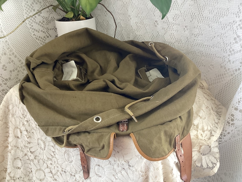 May include: A green canvas backpack with brown leather straps and a drawstring closure. The backpack is open and the inside is visible.