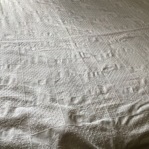 May include: White quilted bedspread with a repeating pattern of squares, each containing a floral or dotted design. The bedspread has a textured surface and a soft, inviting appearance, perfect for a bedroom.