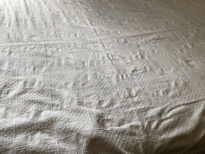 May include: White quilted bedspread with a repeating pattern of squares, each containing a floral or dotted design. The bedspread has a textured surface and a soft, inviting appearance, perfect for a bedroom.