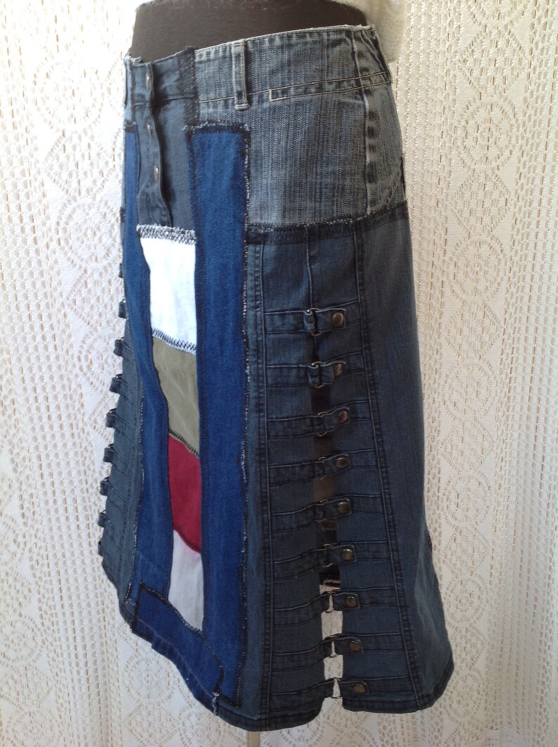 K&ouml;nnte beinhalten: Ein langer Jeansrock aus verschiedenen zusammengen&auml;hten Jeansst&uuml;cken. Der Rock hat eine dunkelblaue Jeansbasis mit einem hellblauen Jeanspanel an der Seite. Der Rock hat eine Reihe von Metallschnallen an der Seite.
