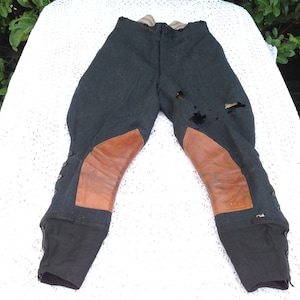Puede incluir: Pantalones de montar gris oscuro con rodilleras de cuero marrón. Los pantalones tienen detalles de botones en la parte inferior de las piernas y muestran signos de desgaste. Los pantalones se exhiben sobre una superficie blanca de ganchillo.