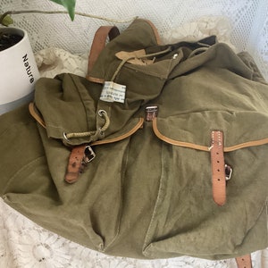 May include: A vintage green canvas backpack with brown leather straps and buckles. The backpack has a drawstring closure and a flap with a buckle.