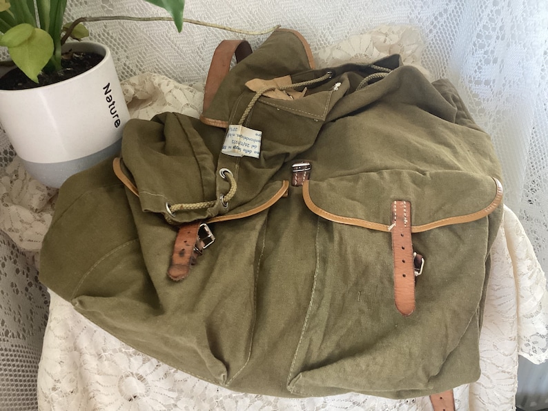 May include: A vintage green canvas backpack with brown leather straps and buckles. The backpack has a drawstring closure and a flap with a buckle.