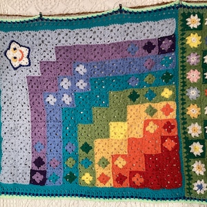 Peut inclure: Une couverture en crochet colorée avec un motif arc-en-ciel et un nuage souriant dans le coin. La couverture a une bordure verte avec des fleurs blanches.