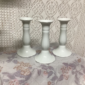 May include: Three white ceramic candle holders with a classic design. Each holder has a wide base, a slender stem, and a flared top. The candle holders are arranged on a floral patterned fabric.