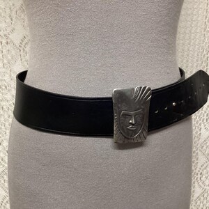 May include: A black leather belt with a silver-colored rectangular buckle. The buckle features a stylized face design. The belt is wide and has several holes for adjustment. The belt is shown on a mannequin.