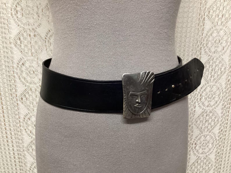 May include: A black leather belt with a silver-colored rectangular buckle. The buckle features a stylized face design. The belt is wide and has several holes for adjustment. The belt is shown on a mannequin.
