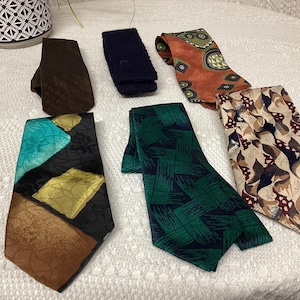 May include: A collection of six neckties in various patterns and colors. The ties include a brown textured tie, a navy blue knit tie, an orange tie with green and black geometric shapes, a green tie with a dark blue brushstroke design, a tie with a black background and teal, gold, and brown shapes, and a tie with a beige background and a pattern of brown, black, and white shapes.