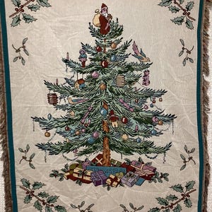 Op de afbeelding: Een wandkleed met een kerstboom met groene, bruine en rode ornamenten. De boom is omringd door cadeaus en een klein kerstmanfiguur. Het wandkleed heeft een witte achtergrond en een turquoise franje rand.